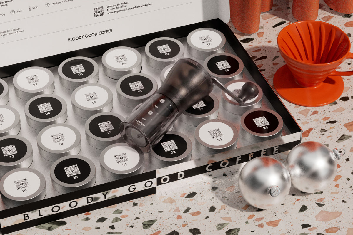 19grams Kaffee Adventskalender 2026 – geöffnete Kalenderbox mit 24 nummerierten silbernen Kaffeedosen mit QR-Code, Handmühle, oranger V60-Handfilter und silberne Weihnachtskugeln auf Terrazzo, Kaffeerösterei Berlin