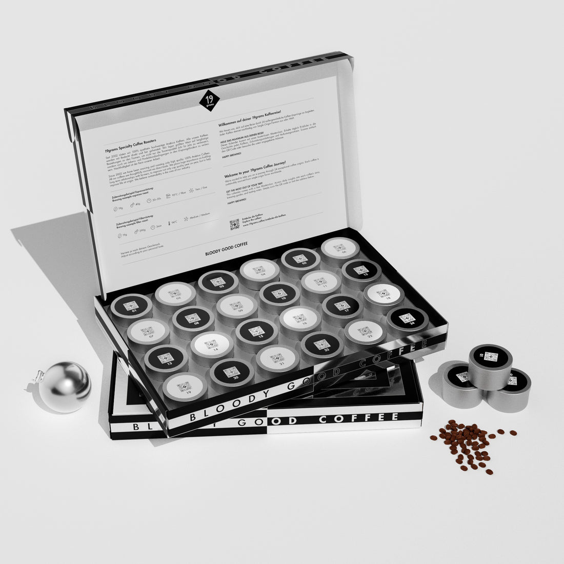 19grams Xmas in July Specialty Coffee Adventskalender – Geöffnete Geschenkbox mit 24 nummerierten Kaffeedosen mit QR-Code, bedrucktem Innendeckel und Schriftzug „Bloody Good Coffee“, mit silberner Weihnachtskugel und Kaffeebohnen auf weißem Hintergrund, Kaffeerösterei Berlin