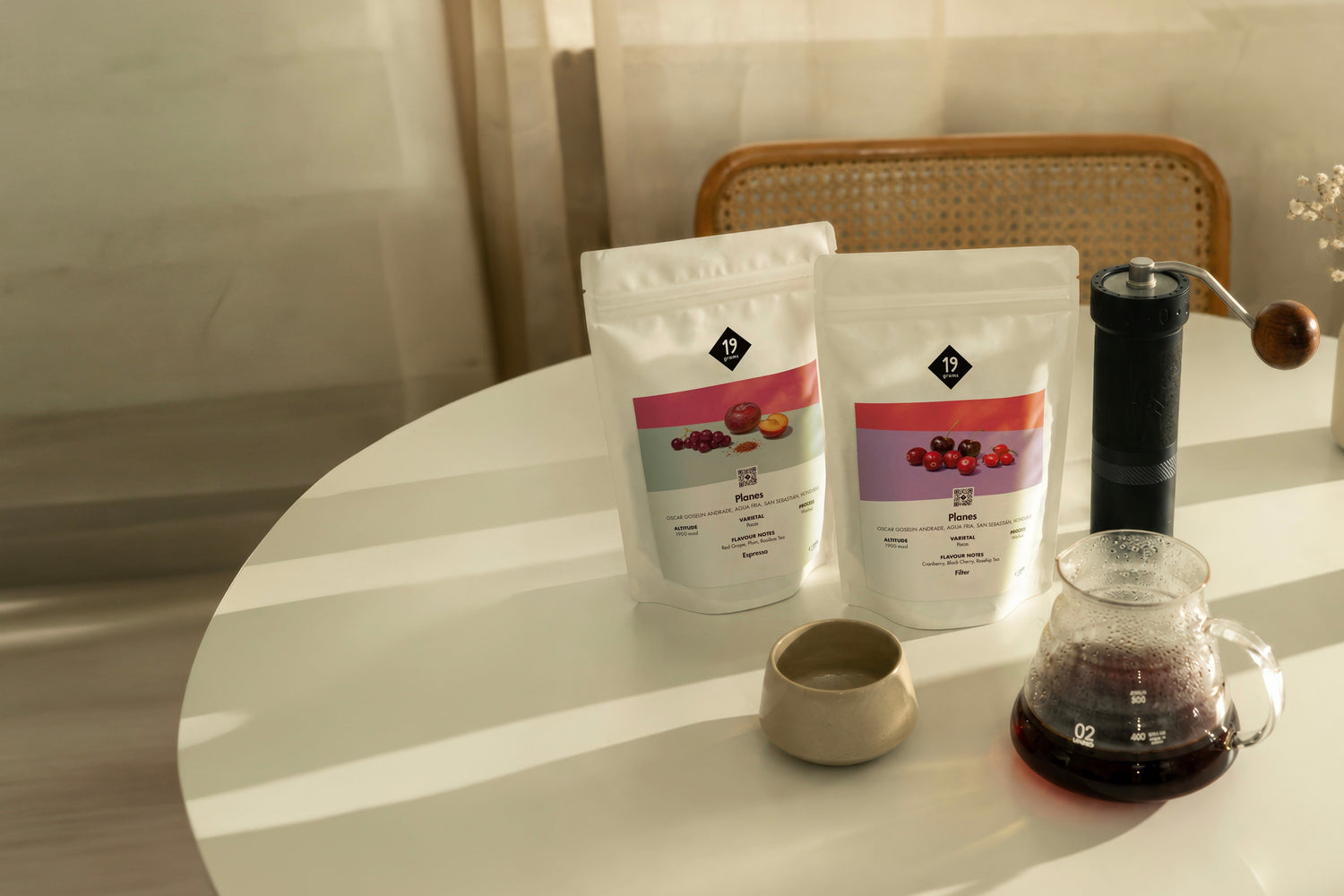 19grams Planes Specialty Coffee Moodbild – Zwei Kaffeebeutel Espresso und Filter auf weißem Tisch mit Handmühle, Keramiktasse und Glaskaraffe mit Filterkaffee im warmen Licht, Single Origin aus Honduras, Kaffeerösterei Berlin