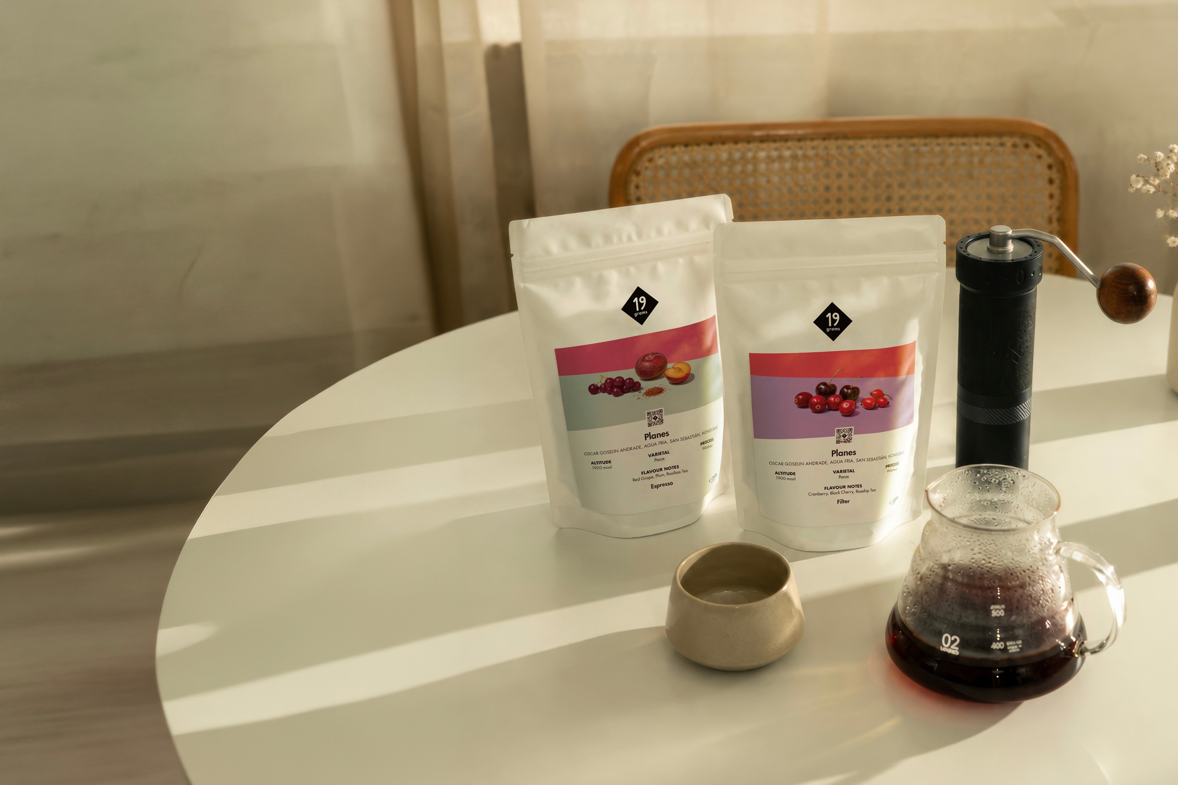 19grams Planes Specialty Coffee Moodbild – Zwei Kaffeebeutel Espresso und Filter auf weißem Tisch mit Handmühle, Keramiktasse und Glaskaraffe mit Filterkaffee im warmen Licht, Single Origin aus Honduras, Kaffeerösterei Berlin
