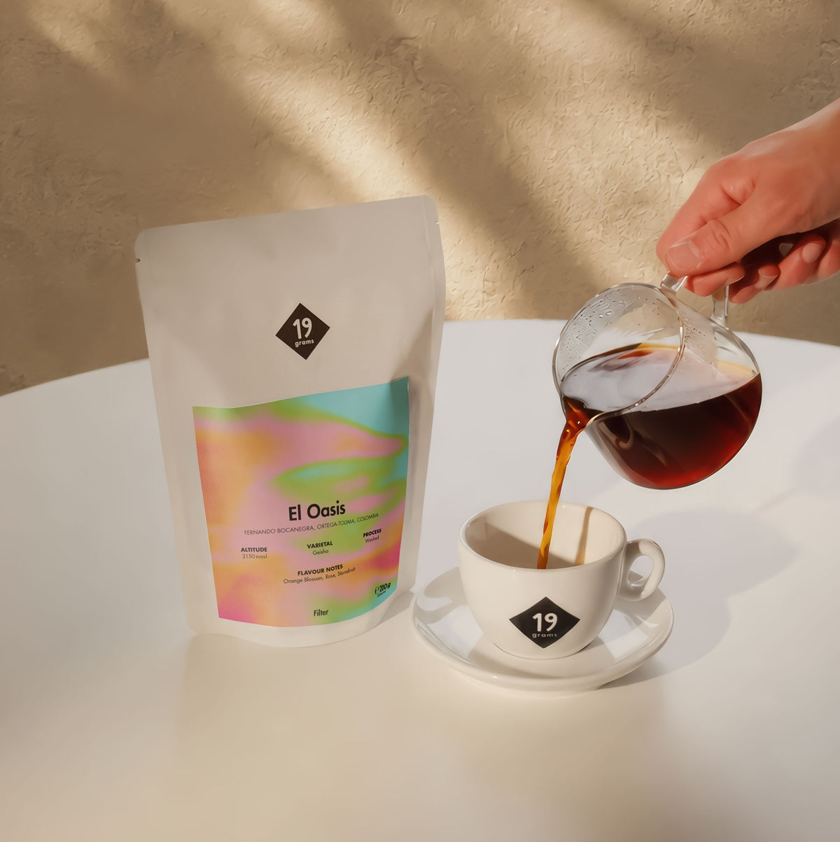 19grams El Oasis Filter – Moodbild mit Kaffeebeutel auf weißem Tisch, während Filterkaffee aus einer Glaskaraffe in eine 19grams Tasse gegossen wird