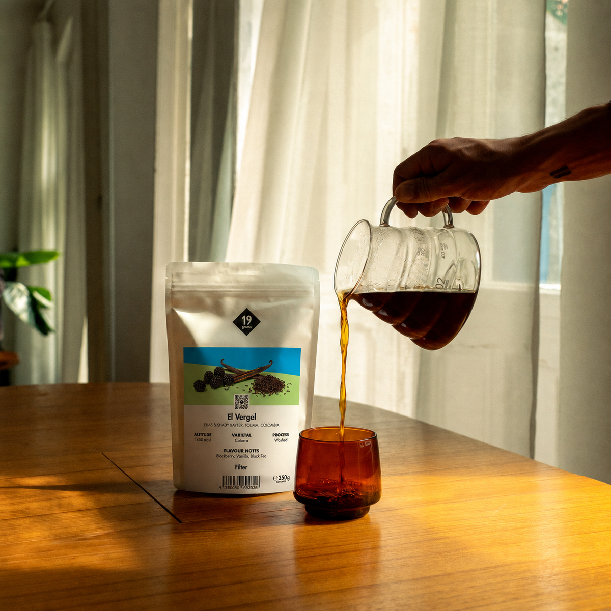 19grams El Vergel Specialty Coffee Filter – Kaffee wird aus Karaffe in Tasse gegossen neben Kaffeebeutel auf Holztisch, Single Origin aus Kolumbien, Kaffeerösterei Berlin