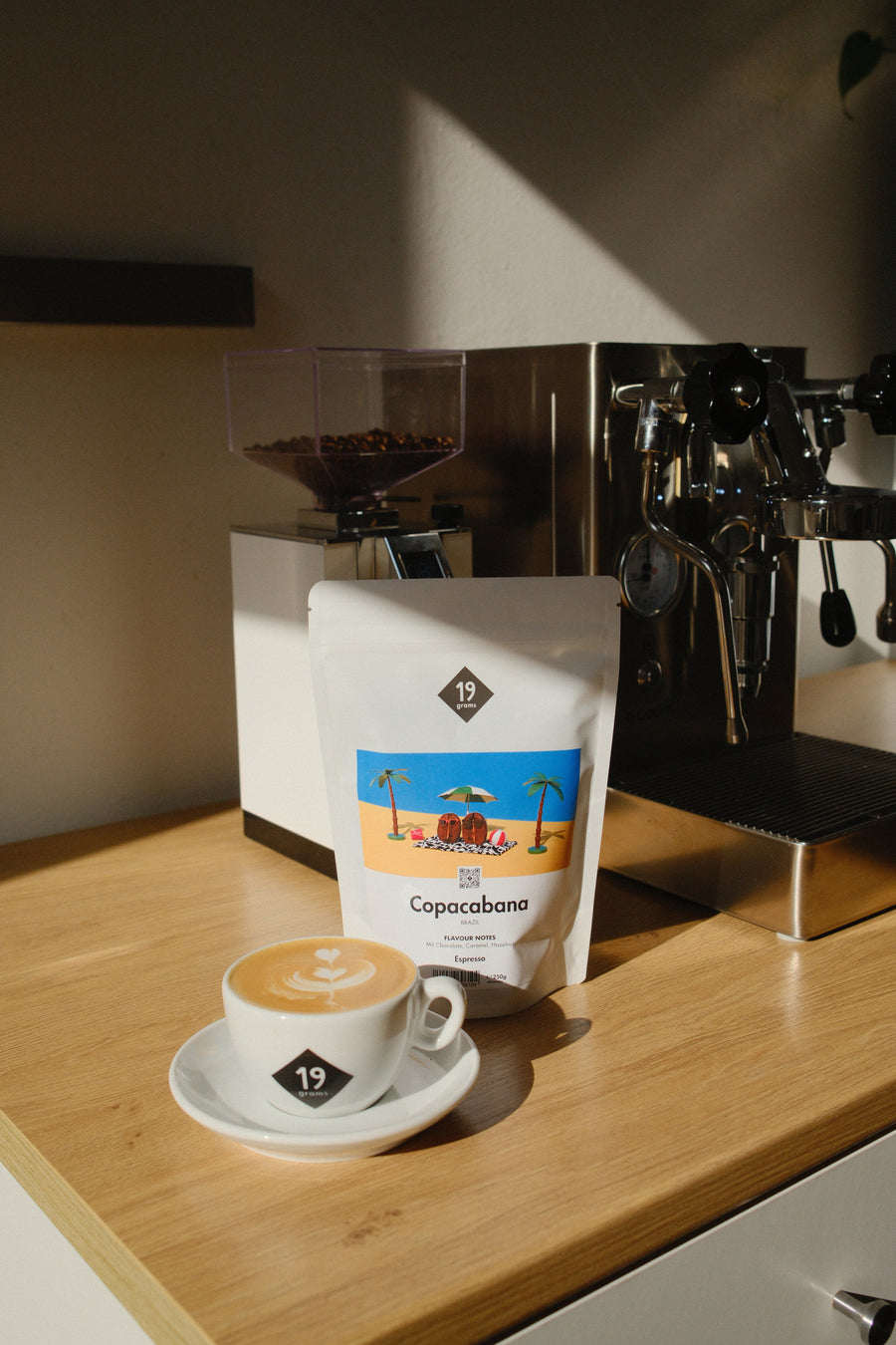 Eine Tüte Copacabana steht neben einer Espressomühle und einer Siebträgermaschine, davor ein Cappuccino mit Latte Art.