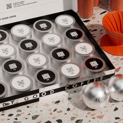 19grams Specialty Coffee Adventskalender 2026 – Geöffneter Kaffee-Adventskalender mit 24 nummerierten Kaffeedosen mit QR-Code, oranger V60-Handfilter und silberne Weihnachtskugel auf Terrazzo-Oberfläche, Kaffeerösterei Berlin