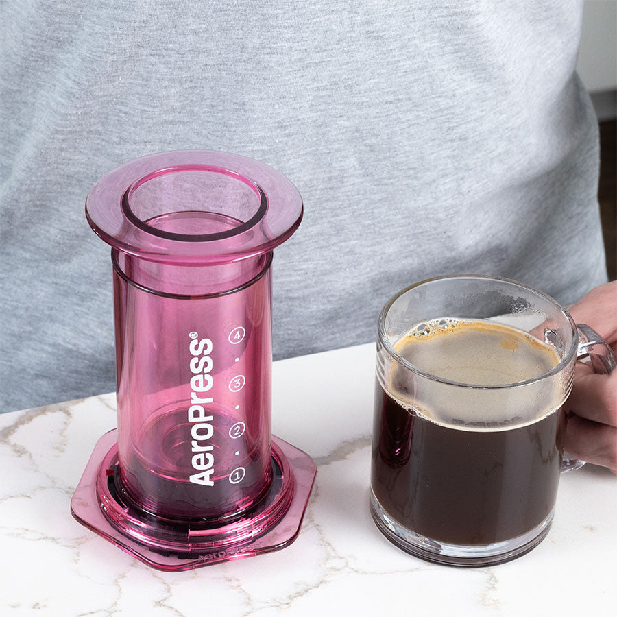 Aeropress Clear Pink neben einer frischen Tasse Kaffee