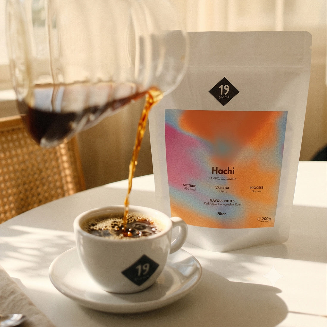 19grams Hachi Filter – Kaffee wird aus einer Glaskaraffe in eine Tasse gegossen, daneben steht der Kaffeebeutel mit buntem Verlaufslabel