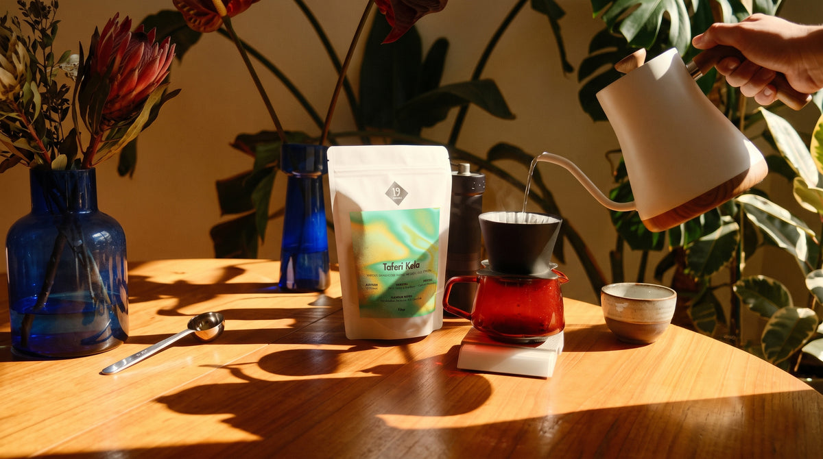 19grams Taferi Kela Filter – Moodbild mit Kaffeebeutel auf einem Holztisch, daneben Handfilter auf roter Glaskaraffe, Wasserkessel, Dosierlöffel und blaue Vasen