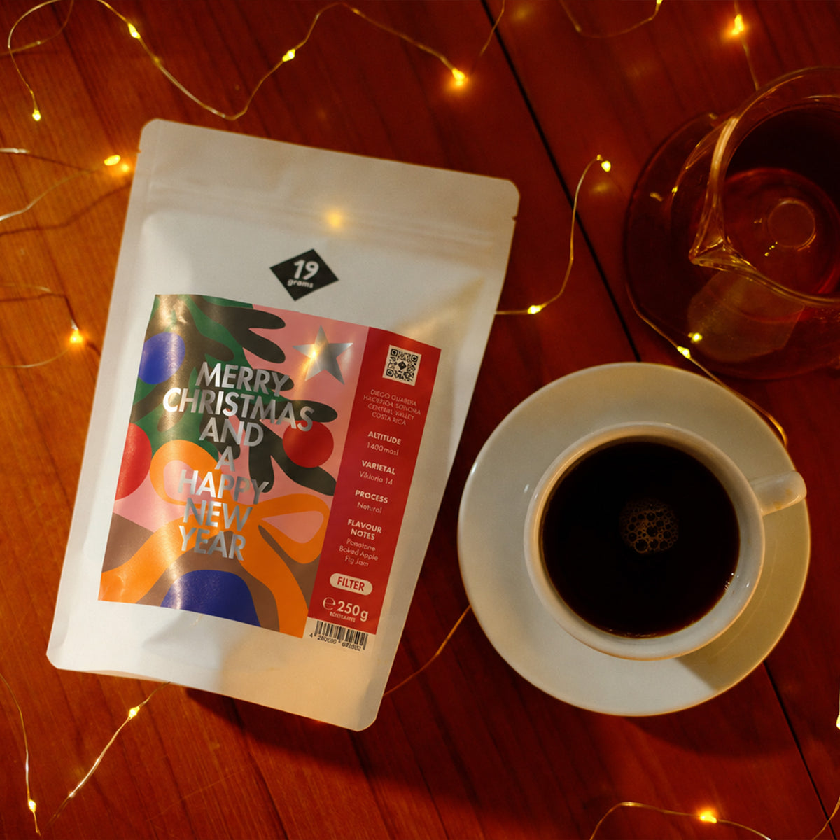 19grams Christmas Coffee Filter – Kaffeebeutel mit weihnachtlicher Illustration auf einem Holztisch, daneben eine Tasse Filterkaffee und eine Glaskaraffe zwischen Lichterketten