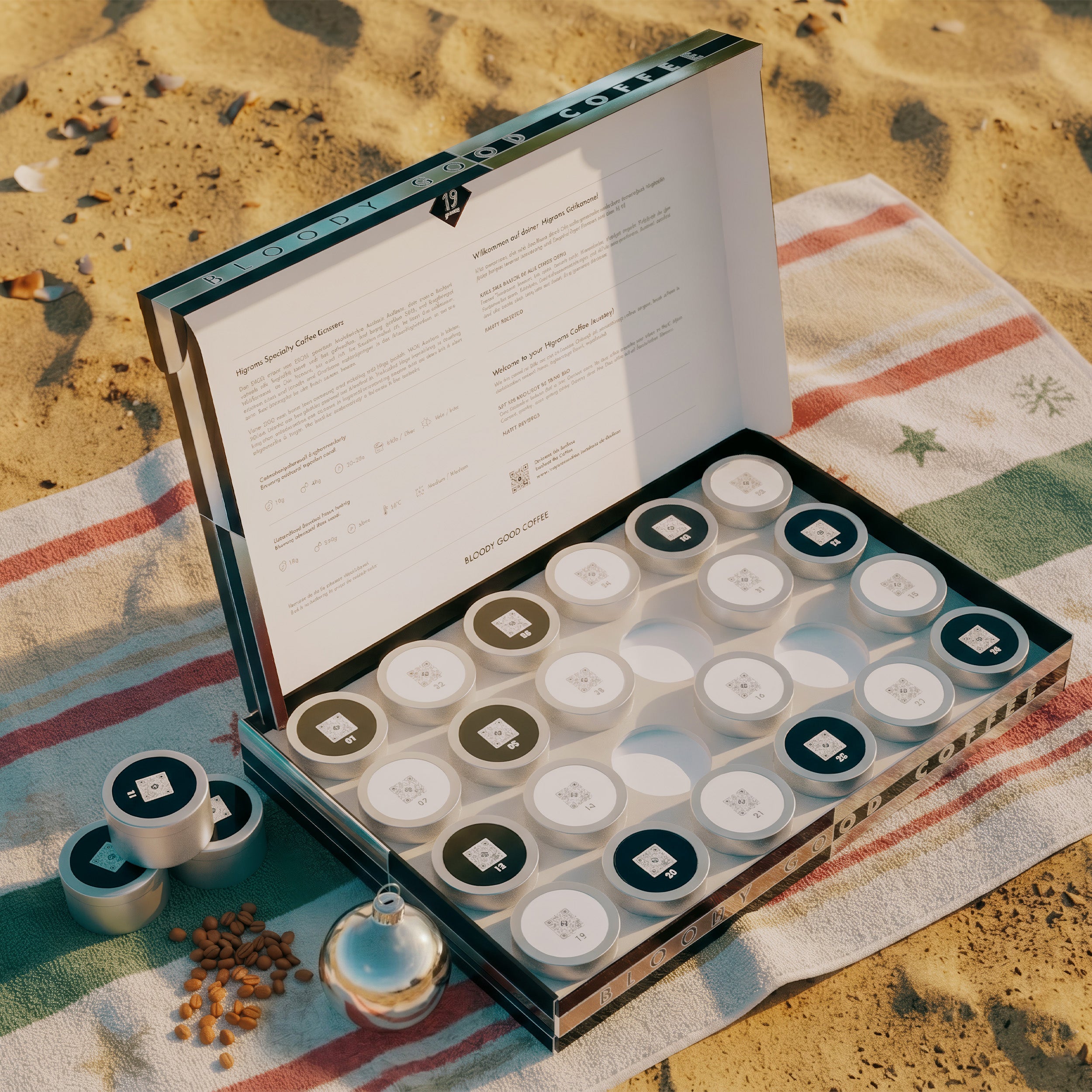 19grams Xmas in July Specialty Coffee Box geöffnet – Sommerliches Moodbild am Strand mit Kaffeedosen, Kaffeebohnen und Christbaumkugel auf Handtuch, Kaffeerösterei Berlin