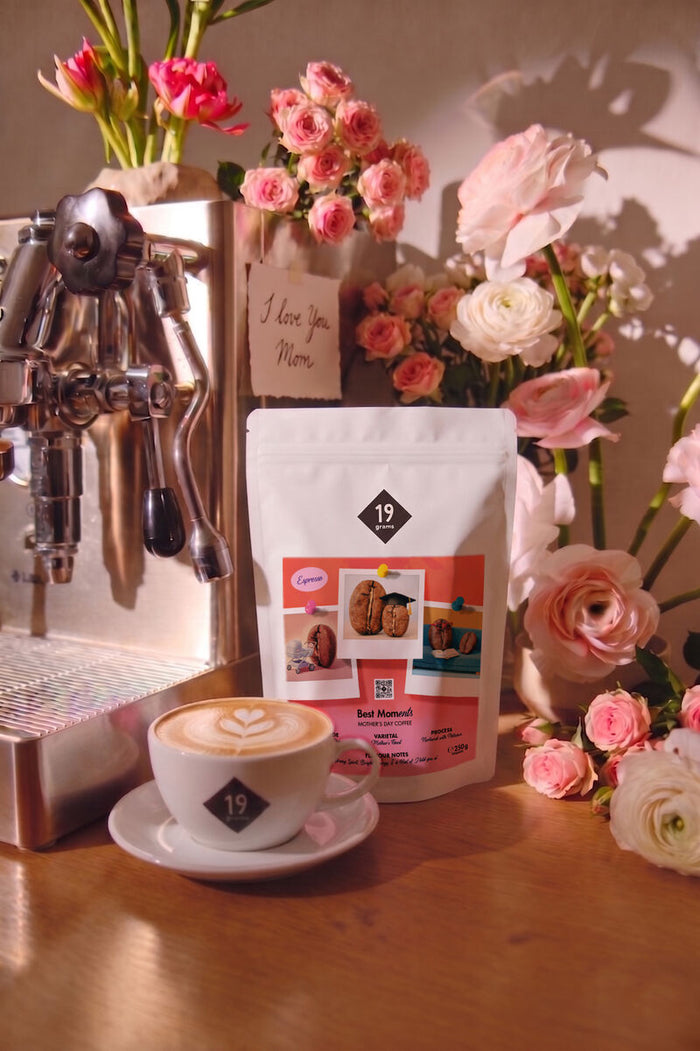 Cappuccino mit Latte-Art-Herz vor einer Siebträgermaschine, daneben die Best-MOMents-Espressotüte und rosa Rosen
