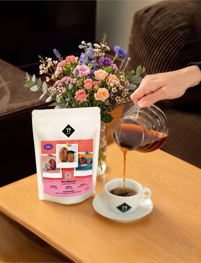 Kaffee läuft aus einer Glaskaraffe in eine 19grams-Tasse, daneben die Best-MOMents-Filtertüte und ein Blumenstrauß