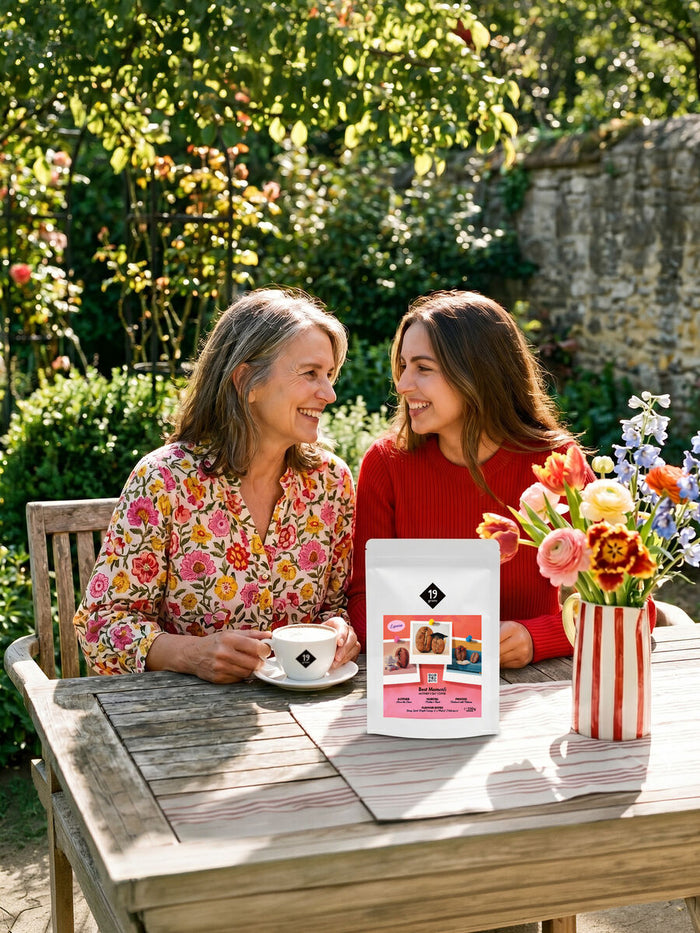 Mutter und Tochter lachen sich im Garten an, auf dem Holztisch Kaffee und die Best-MOMents-Tüte
