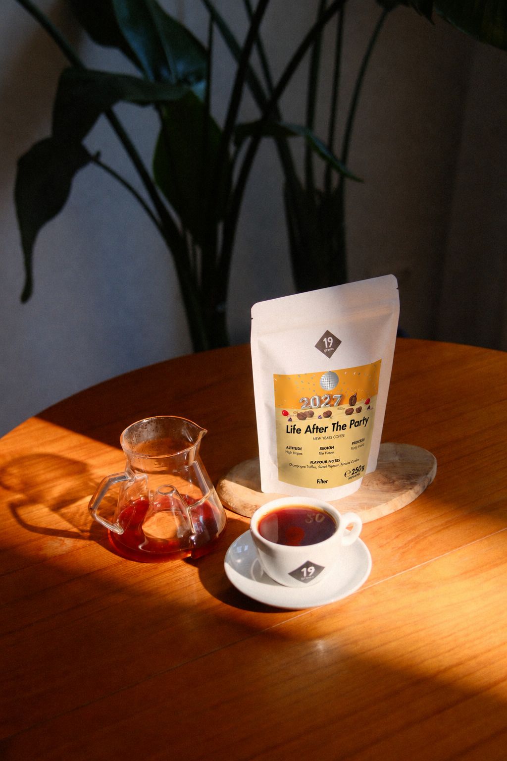 19grams Life After The Party Kaffee – 250 g – Kaffeebeutel mit goldenem Neujahrslabel, Glaskanne und volle Tasse auf einem Holztisch im Sonnenlicht